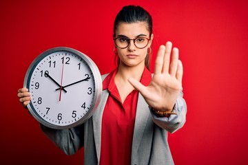 Young beautiful brunette businesswoman doing countdown holding big clock with open hand doing stop...