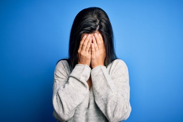 Young beautiful brunette woman wearing casual sweater standing over blue background with sad expression covering face with hands while crying. Depression concept.