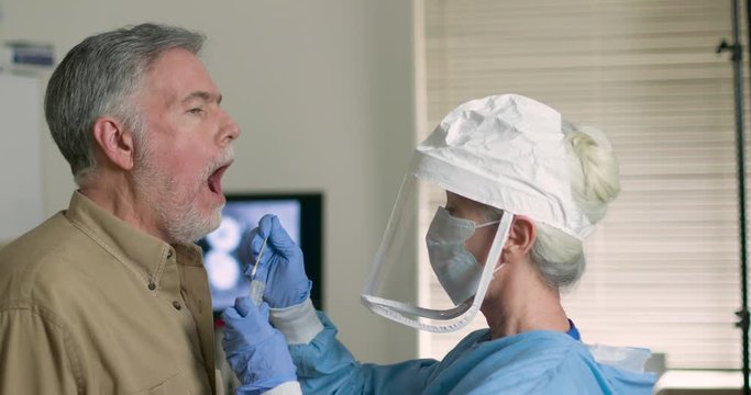 A mature Caucasian man in a clinical setting being swabbed by a technician in protective garb to determine if he has contracted the coronavirus or COVID-19.