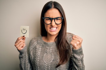 Young beautiful brunette woman wearing glasses holding paper with smile emoji annoyed and frustrated shouting with anger, crazy and yelling with raised hand, anger concept