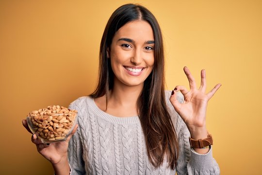 Young beautiful brunette woman holding bowl with peanuts over isolated yellow background doing ok sign with fingers, excellent symbol