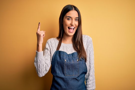 Young beautiful baker woman wearing apron uniform cooking over yellow background pointing finger up with successful idea. Exited and happy. Number one.
