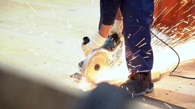 Unrecognizable worker cuts metal rods into bars on construction site. Man hold, use grinder, sawing steel rebar on floor on wooden sheets. Guy does his job, sparks fly on factory area. Indoor close up