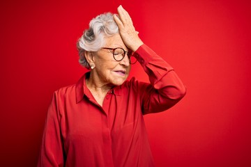 Senior beautiful grey-haired woman wearing casual shirt and glasses over red background surprised...