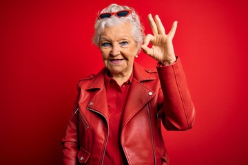 Senior beautiful grey-haired woman wearing casual red jacket and sunglasses smiling positive doing ok sign with hand and fingers. Successful expression.