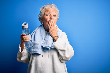 Senior beautiful sporty woman holding bottle of water standing over isolated blue background cover mouth with hand shocked with shame for mistake, expression of fear, scared in silence, secret concept