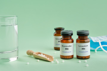 Bottles of homeopathic drugs - Bryonia, Lycopodium, Gelsemium - recommended by homeopaths for treatment and prophylaxis of Coronavirus Covid-19, glass of water, scoop, medical protective mask.