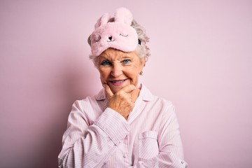 Senior beautiful woman wearing sleep mask and pajama over isolated pink background looking confident at the camera smiling with crossed arms and hand raised on chin. Thinking positive.