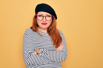 Beautiful redhead woman wearing striped t-shirt and french beret over yellow background skeptic and...