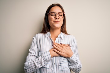 Young beautiful brunette woman wearing casual shirt and glasses over white background smiling with hands on chest with closed eyes and grateful gesture on face. Health concept.