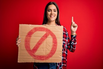 Young beautiful brunette woman holding banner with prohibited signal over red background surprised with an idea or question pointing finger with happy face, number one