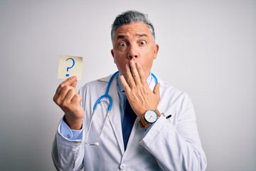 Middle age handsome grey-haired doctor man holding paper with question mark cover mouth with hand shocked with shame for mistake, expression of fear, scared in silence, secret concept