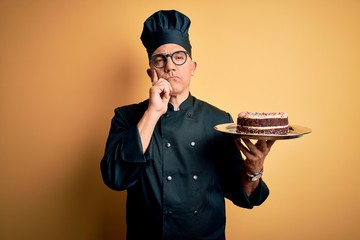 Middle age handsome grey-haired baker man wearing cooker uniform and hat holding cake serious face...