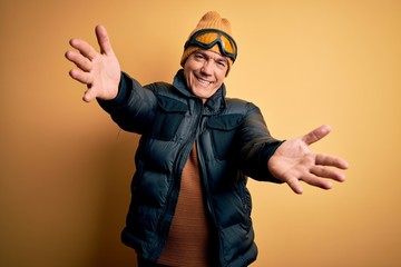 Middle age handsome grey-haired skier man on vacation wearing ski goggles looking at the camera smiling with open arms for hug. Cheerful expression embracing happiness.