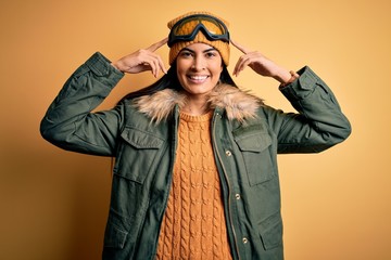 Young beautiful hispanic woman wearing ski glasses and coat for winter weather smiling pointing to head with both hands finger, great idea or thought, good memory