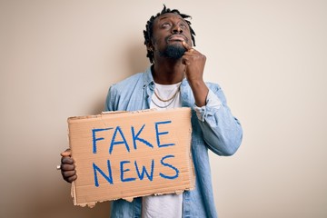 Young african american man holding banner with fake news message over white background serious face...