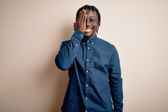 Young handsome african american man wearing casual shirt standing over white background covering one eye with hand, confident smile on face and surprise emotion.