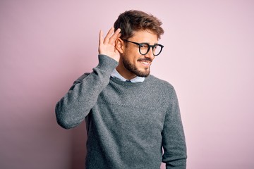 Young handsome man with beard wearing glasses and sweater standing over pink background smiling with hand over ear listening an hearing to rumor or gossip. Deafness concept.