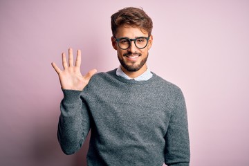Young handsome man with beard wearing glasses and sweater standing over pink background showing and...
