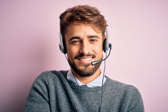 Young call center agent man with beard wearing headset over isolated pink background happy face smiling with crossed arms looking at the camera. Positive person.