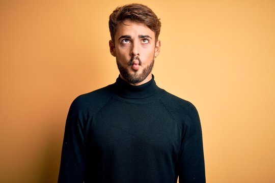 Young handsome man with beard wearing turtleneck sweater standing over yellow background making fish face with lips, crazy and comical gesture. Funny expression.