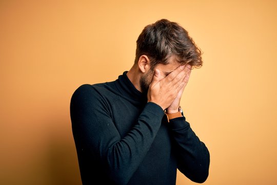Young handsome man with beard wearing turtleneck sweater standing over yellow background with sad expression covering face with hands while crying. Depression concept.