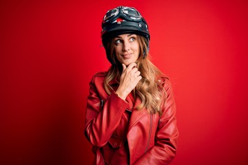 Young beautiful brunette motrocyclist woman wearing moto helmet over red background with hand on chin thinking about question, pensive expression. Smiling with thoughtful face. Doubt concept.