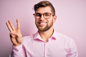 Young handsome blond man with beard and blue eyes wearing pink shirt and glasses showing and...