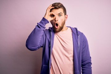 Young blond man with beard and blue eyes wearing purple sweatshirt over pink background doing ok gesture shocked with surprised face, eye looking through fingers. Unbelieving expression.