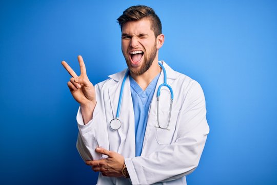 Young blond doctor man with beard and blue eyes wearing white coat and stethoscope smiling with happy face winking at the camera doing victory sign. Number two.