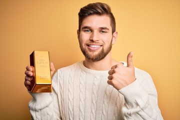 Young blond man with beard and blue eyes holding gold ingot over isolated yellow background happy...