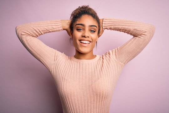 Young beautiful african american girl wearing casual sweater standing over pink background relaxing and stretching, arms and hands behind head and neck smiling happy