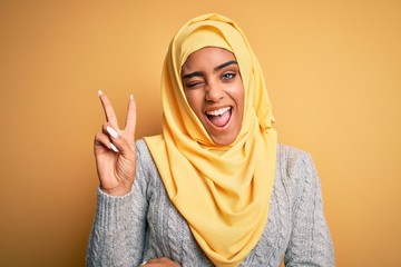 Young beautiful african american girl wearing muslim hijab over isolated yellow background smiling with happy face winking at the camera doing victory sign with fingers. Number two.