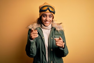 Young african american afro skier girl wearing snow sportswear and ski goggles pointing fingers to camera with happy and funny face. Good energy and vibes.