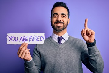 Young handsome businessman with beard holding paper with you are fired message surprised with an idea or question pointing finger with happy face, number one