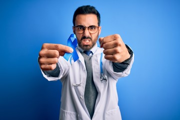 Young handsome doctor man with beard wearing stethoscope holding blue cancer ribbon annoyed and frustrated shouting with anger, crazy and yelling with raised hand, anger concept