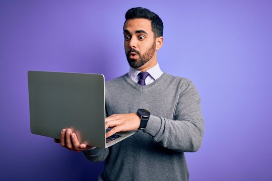Young handsome businessman with beard working using laptop over purple background scared in shock with a surprise face, afraid and excited with fear expression