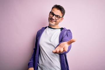 Young handsome man wearing purple sweatshirt and glasses standing over pink background smiling cheerful offering palm hand giving assistance and acceptance.