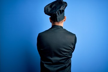 Young handsome chef man wearing cooker uniform and hat over isolated blue background standing...