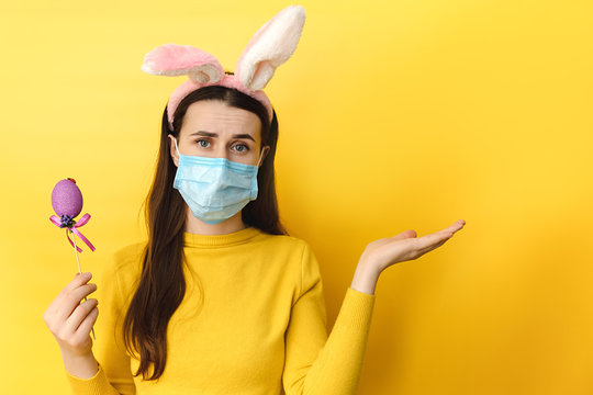 Displeased young woman with virus mask, wears bunny ears, raises palm as if showing something, gestures over blank space, holds easter egg, isolated on yellow wall. Coronavirus and pandemic concept.
