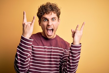 Fototapeta premium Young blond handsome man with curly hair wearing casual striped sweater shouting with crazy expression doing rock symbol with hands up. Music star. Heavy concept.
