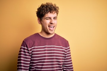 Young blond handsome man with curly hair wearing casual striped sweater winking looking at the...