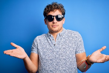 Young blond handsome man with curly hair on vacation wearing striped polo and sunglasses clueless...