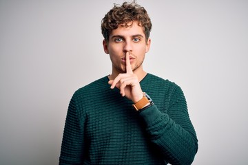 Young blond handsome man with curly hair wearing green sweater over white background asking to be quiet with finger on lips. Silence and secret concept. - obrazy, fototapety, plakaty