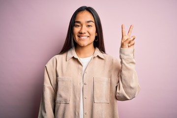 Young beautiful asian woman wearing casual shirt standing over pink background showing and pointing up with fingers number two while smiling confident and happy.