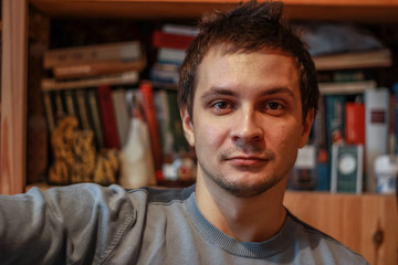 Obraz premium Portrait of a young seriously man at home on the background of the bookcase looking to camera.