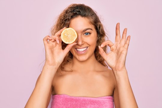 Beautiful woman with blue eyes wearing towel shower after bath holding lemon fruit over eye doing ok sign with fingers, excellent symbol