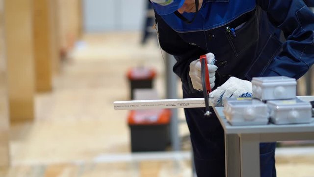 One male worker in overalls, wear white gloves, standing at table, holding hand hacksaw for metal, sawing off piece of plastic cable channel in large workshop room at factory background. Copy space