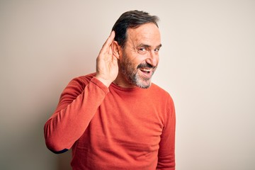 Middle age hoary man wearing casual orange sweater standing over isolated white background smiling with hand over ear listening an hearing to rumor or gossip. Deafness concept.