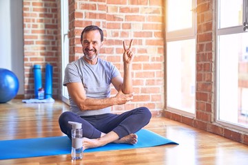 Middle age handsome sportman sitting on mat doing stretching yoga exercise at gym smiling with...
